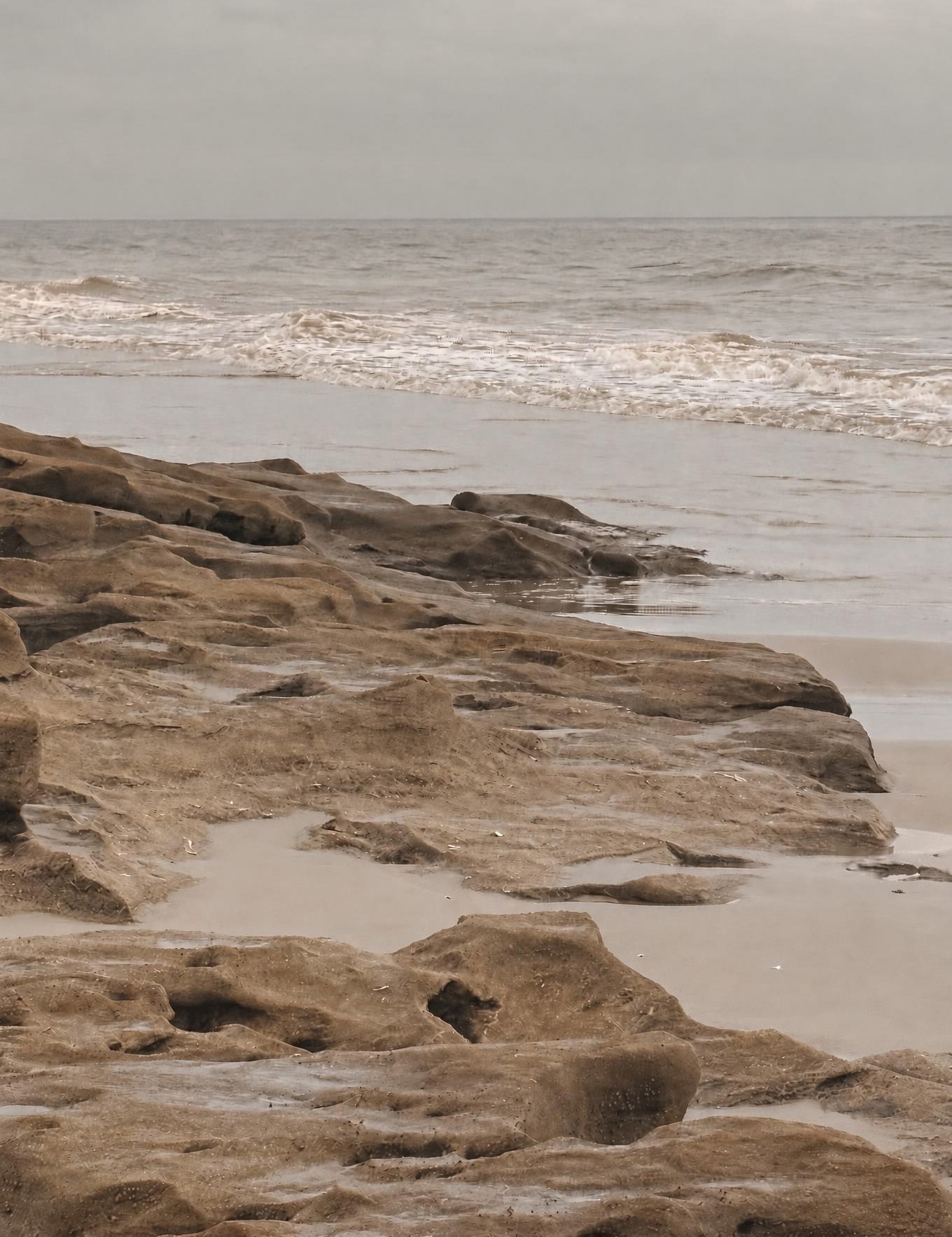 Pitted rock ledge in wet sand with gentle surf under soft, warm light